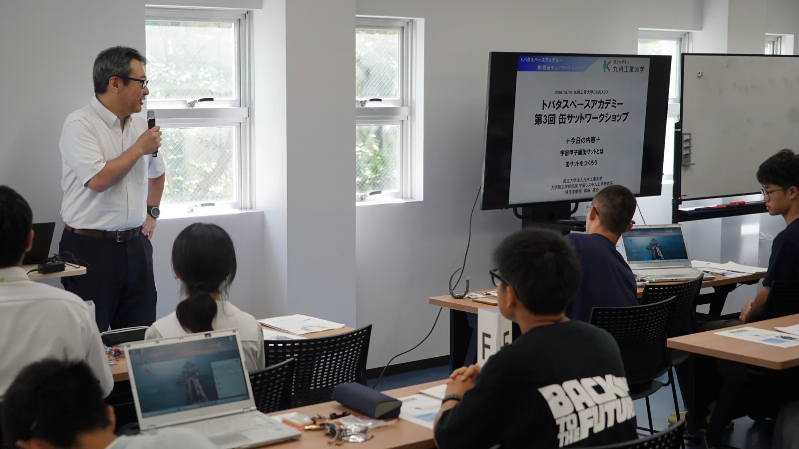 「トバタスペースアカデミー 第3回 缶サットワークショップ」のタイトルスライドを背にマイクで話す前田恵介先生